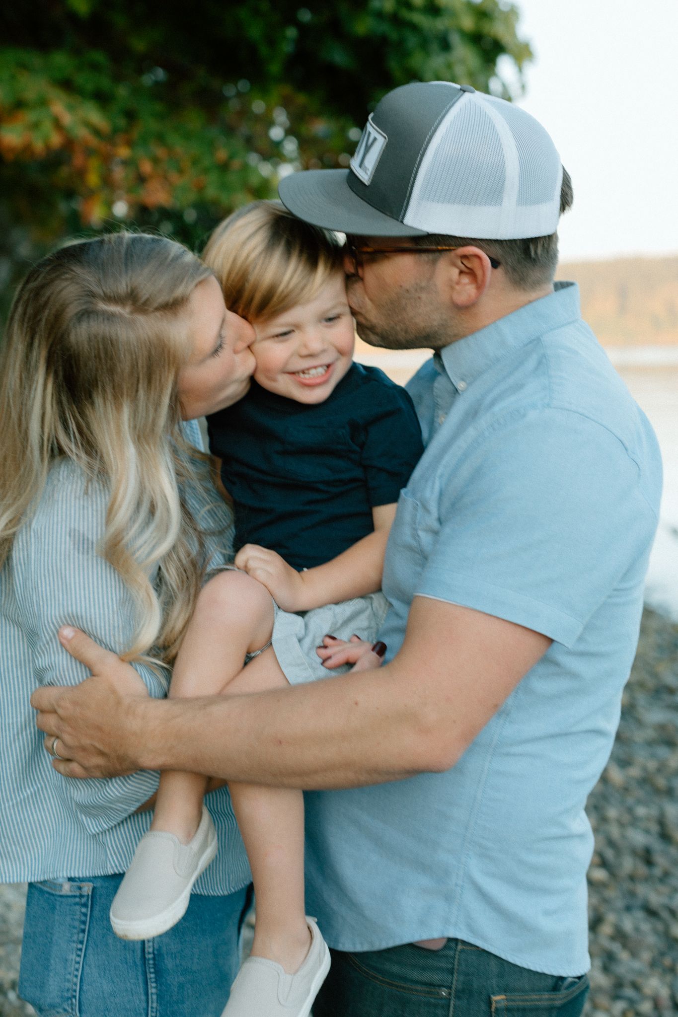 Parents in blue outfits kissing their smiling toddler son on the cheeks outdoors near a rocky shore.