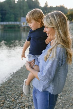 Blonde mother in striped blue shirt holding her young son near a peaceful waterfront at golden hour.