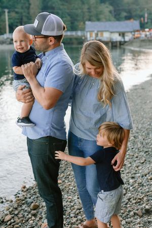 Happy family of four with two young boys standing on a rocky beach near calm water at sunset.