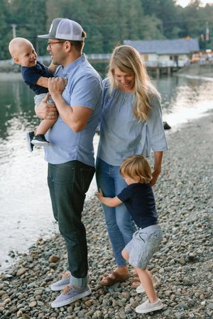 Family of four enjoying a candid moment on a pebble beach, father holding baby while toddler hugs mom.