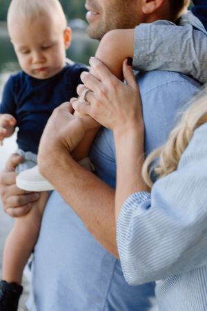 Close-up of mother wearing wedding ring embracing father holding baby in navy outfit outdoors.