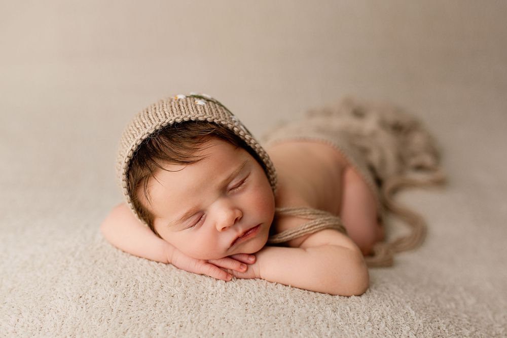 Peaceful newborn baby wearing knit bonnet in hands under chin pose on cream blanket at Summerville SC studio