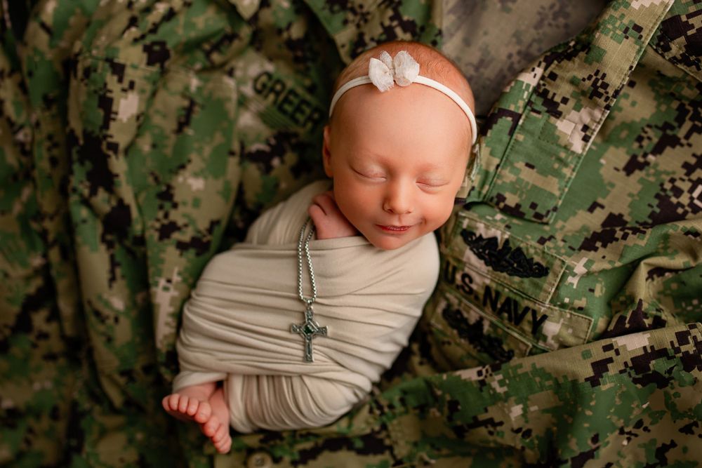 Newborn baby in beige wrap with white flower headband on military camouflage uniform backdrop in Summerville SC