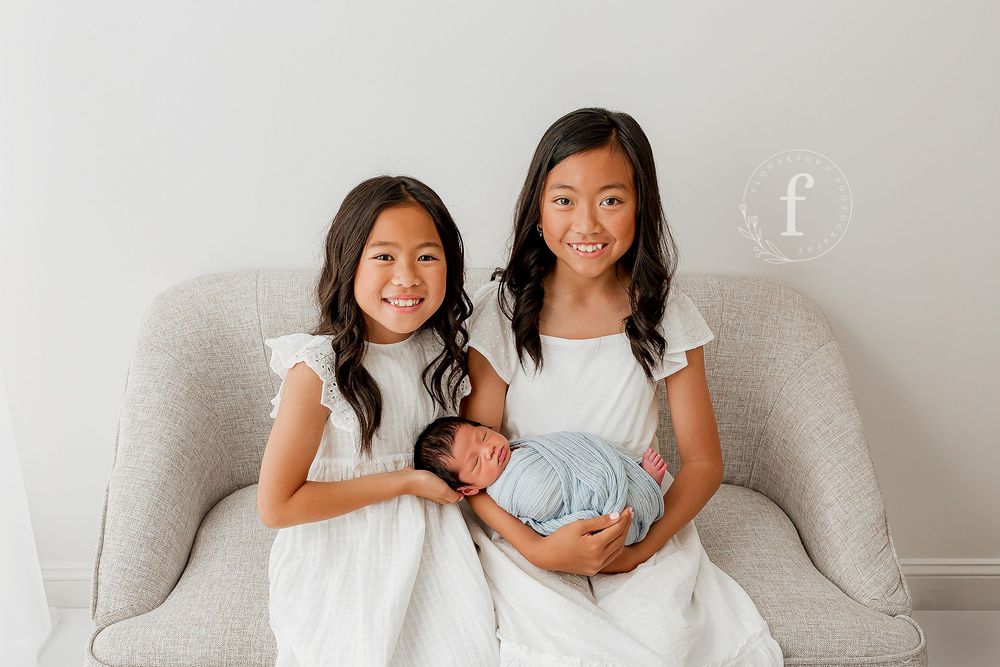 Two sisters in white dresses holding newborn baby brother on gray chair at Summerville SC photography studio