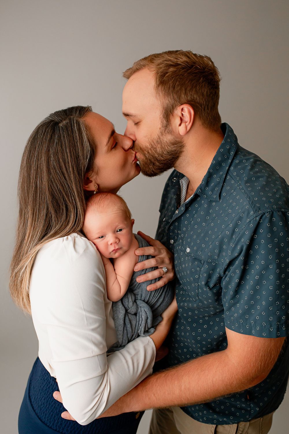 Loving parents kissing while holding newborn baby in gray wrap during family portrait session in Summerville SC