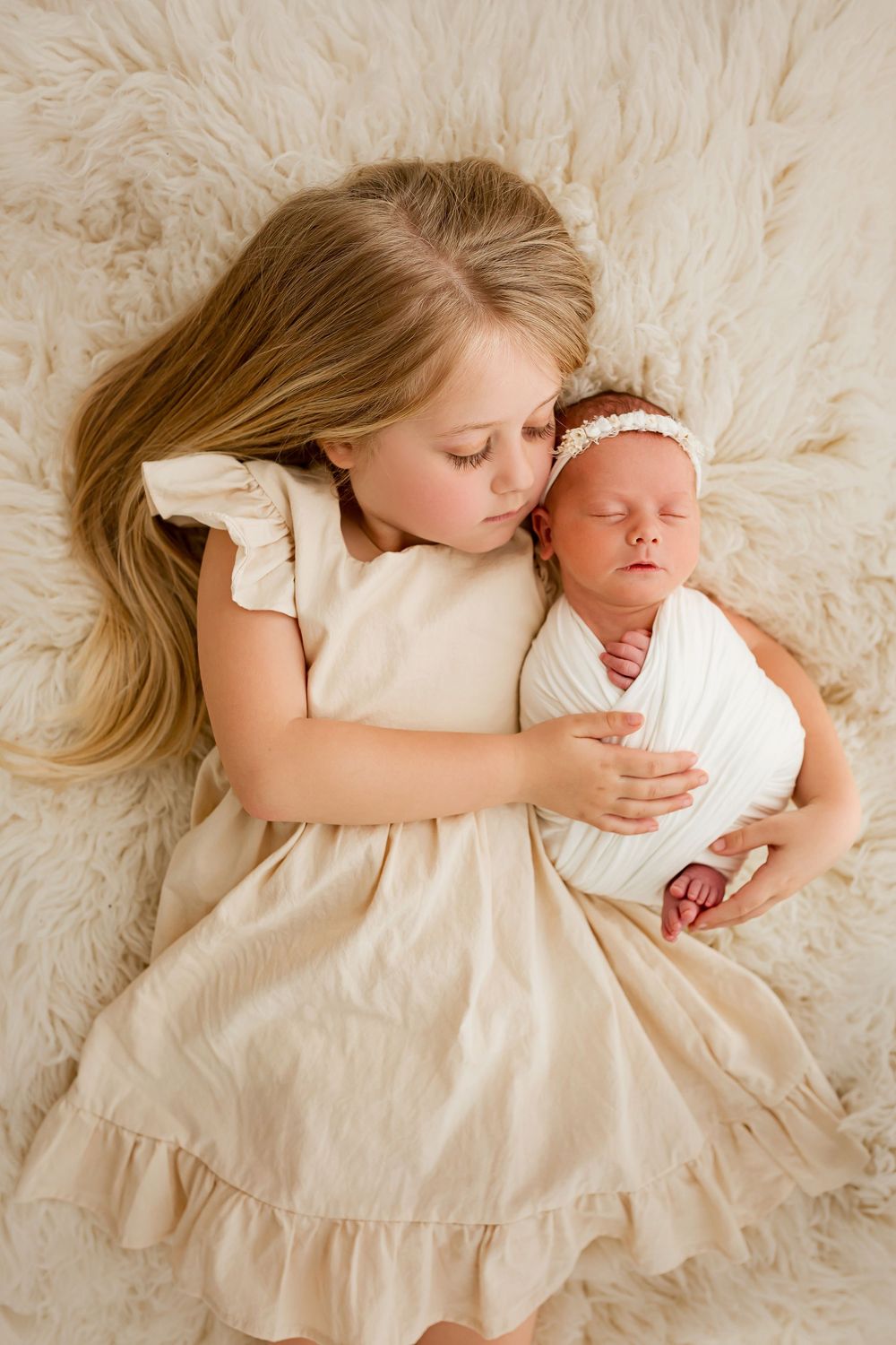 Sweet sibling portrait of big sister holding newborn baby in cream wrap and headband on fluffy blanket