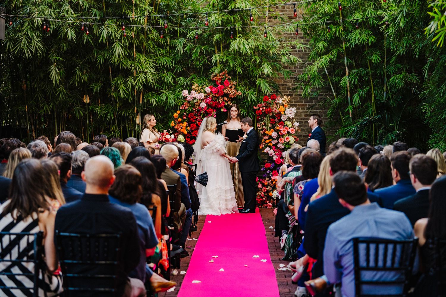 Wedding ceremony on pink carpet aisle with bamboo and red floral arch surrounded by guests in lush garden setting.
