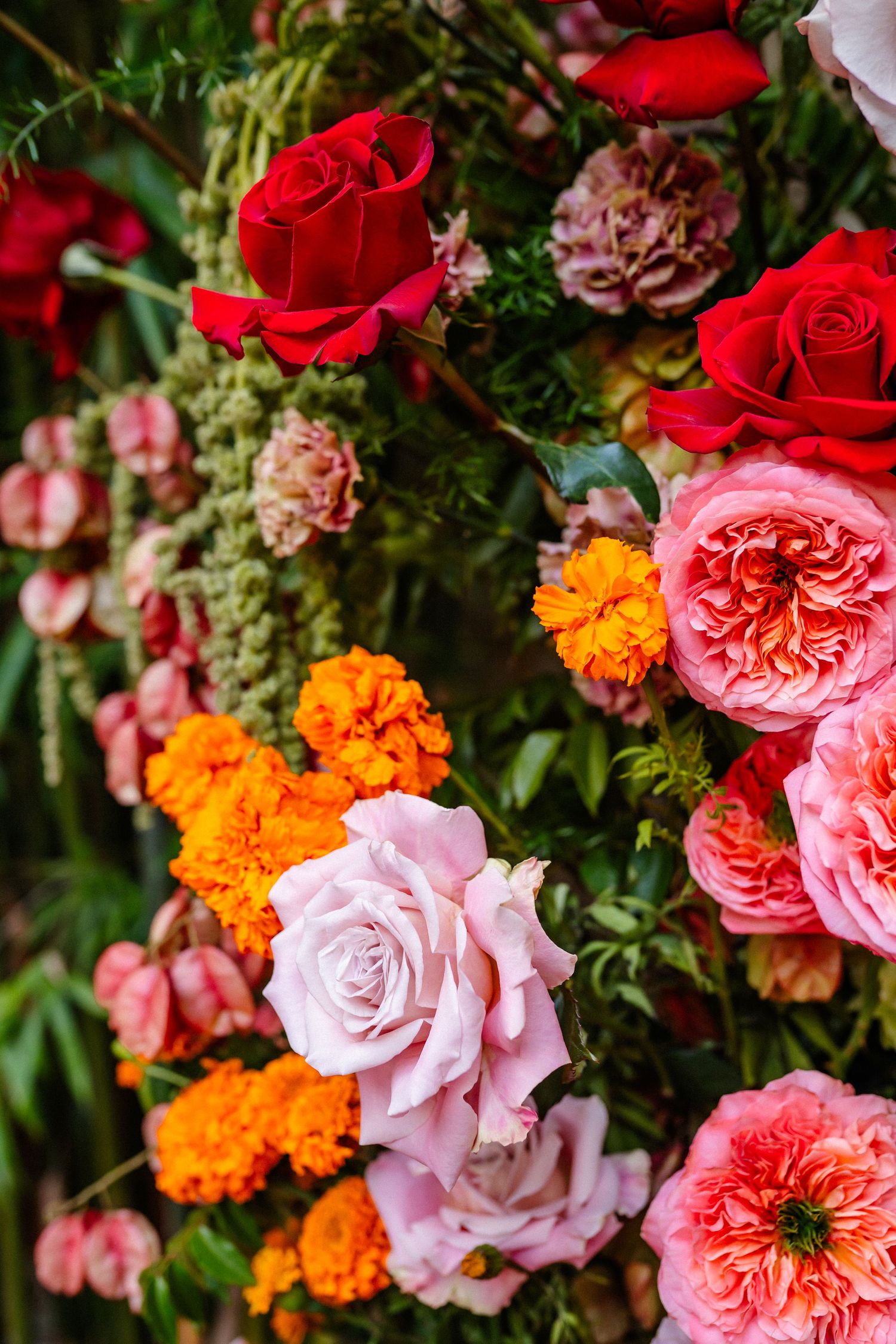 Vibrant floral arrangement featuring red roses, pink garden roses, orange marigolds, and lush greenery in a colorful display.