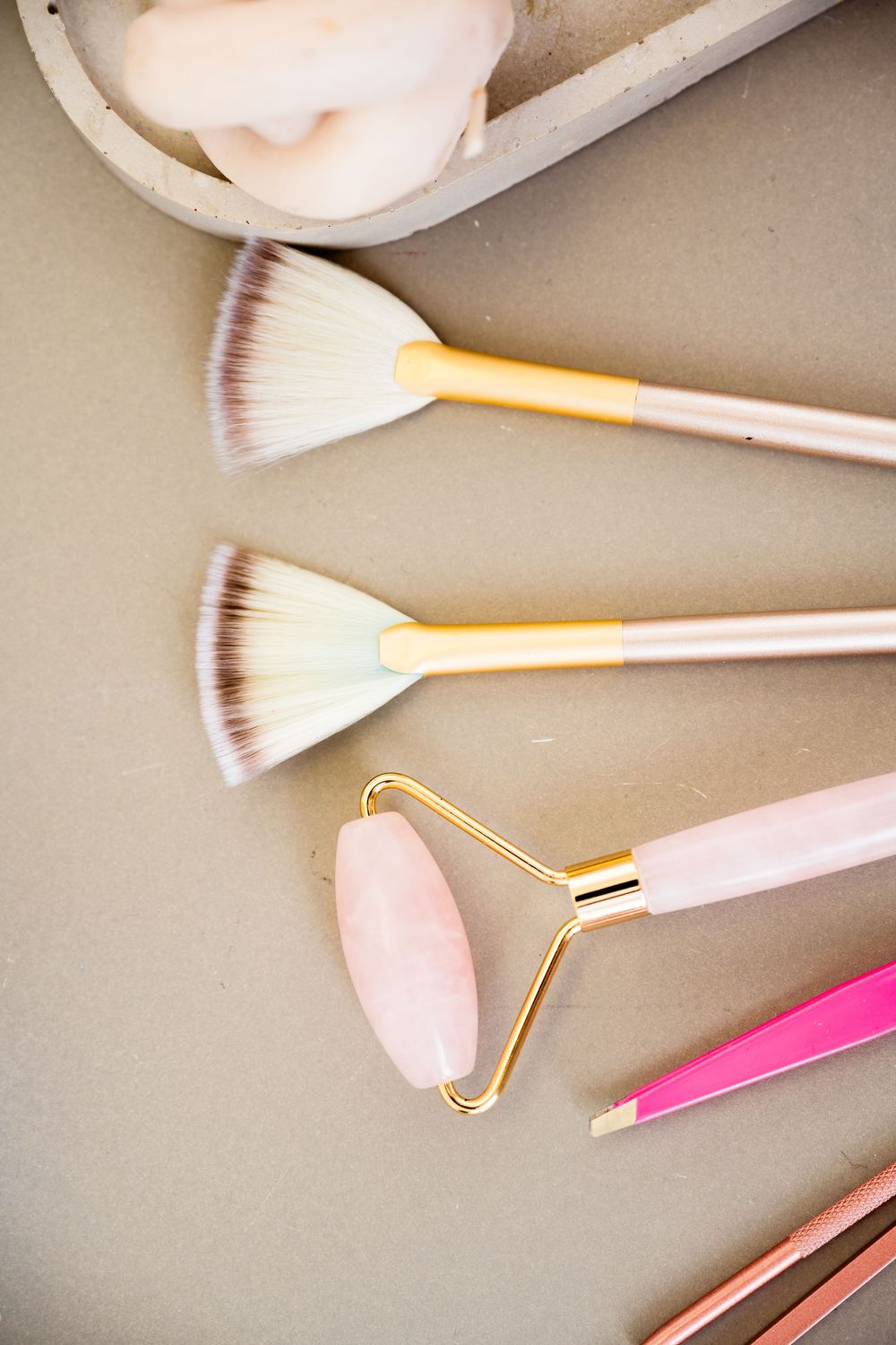 Two fan makeup brushes with gold handles, a pink rose quartz facial roller, and pink tweezers arranged on a taupe surface.