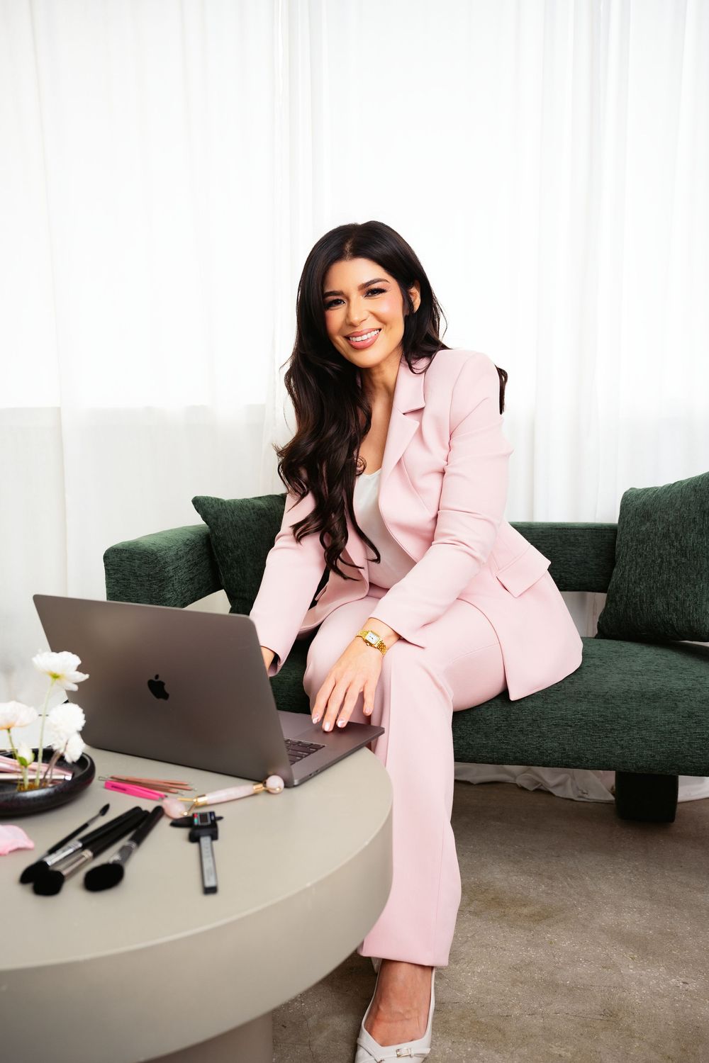Smiling woman in a pink blazer and pants sits on a green sofa beside a laptop, makeup brushes, and white flowers on a round table.