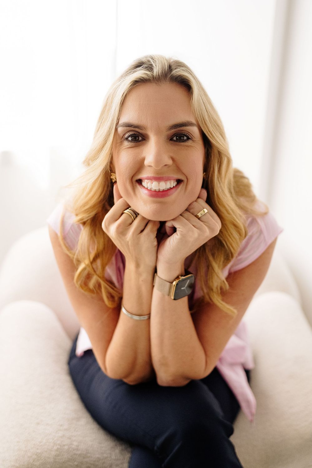 Smiling woman with long wavy blonde hair rests her chin on her hands while seated on a cozy cream armchair in a bright room.