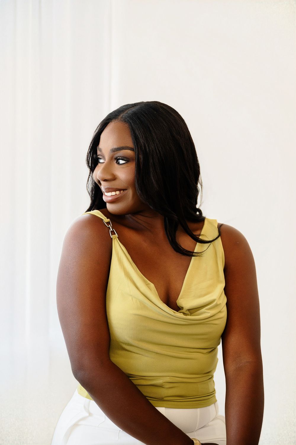 Smiling woman with long dark hair in a yellow cowl-neck satin top and white pants looks over her shoulder against a soft white backdrop.