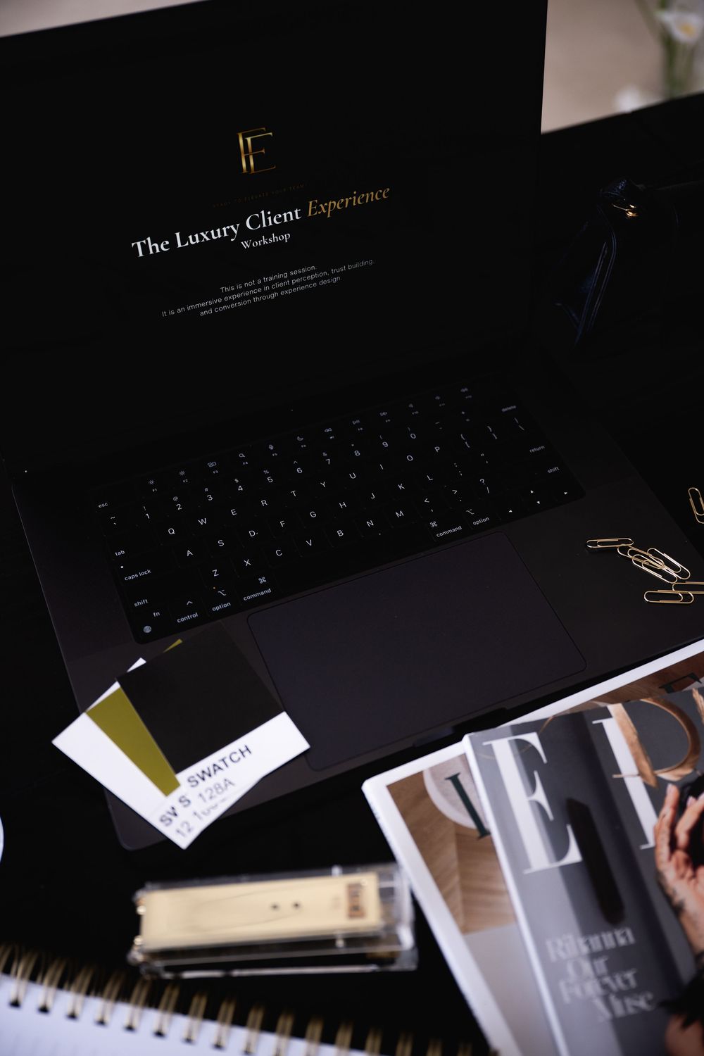 Laptop displaying The Luxury Client Experience Workshop beside a color swatch, paper clips, a notebook, and a magazine on a dark desk.
