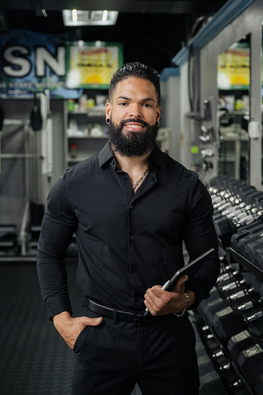 Fitness trainer in black attire stands confidently in gym with dumbbells in background.