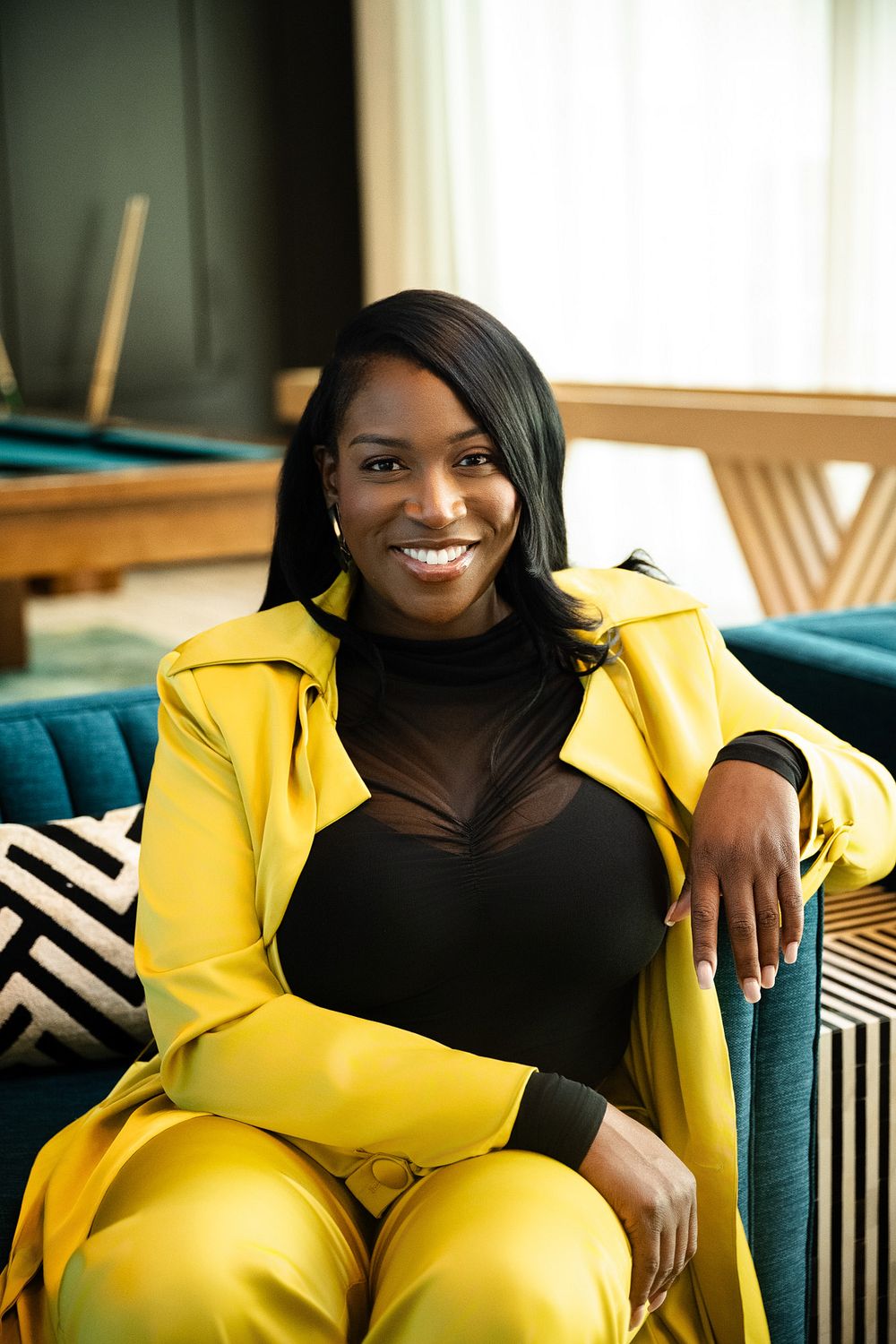 Smiling woman in a yellow blazer and trousers over a black top sits relaxed on a teal sofa in a stylish, sunlit modern room.