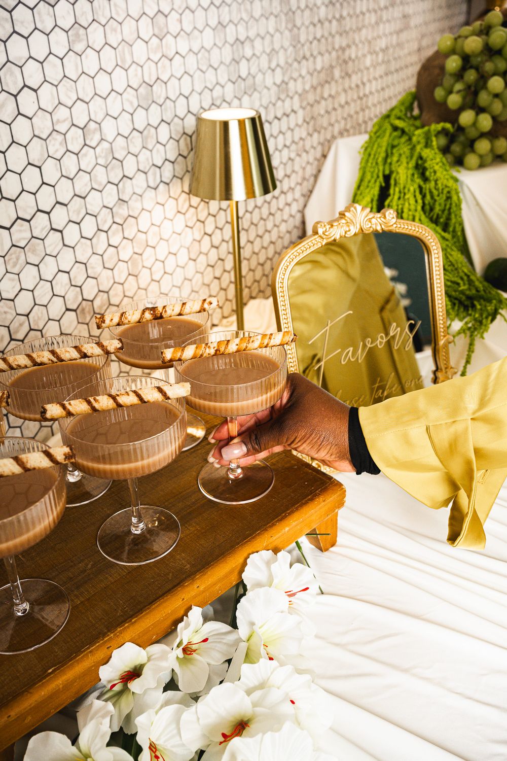 Hand reaching for chocolate espresso martini coupes with wafer garnishes on a wooden table beside white orchids and a gold lamp.
