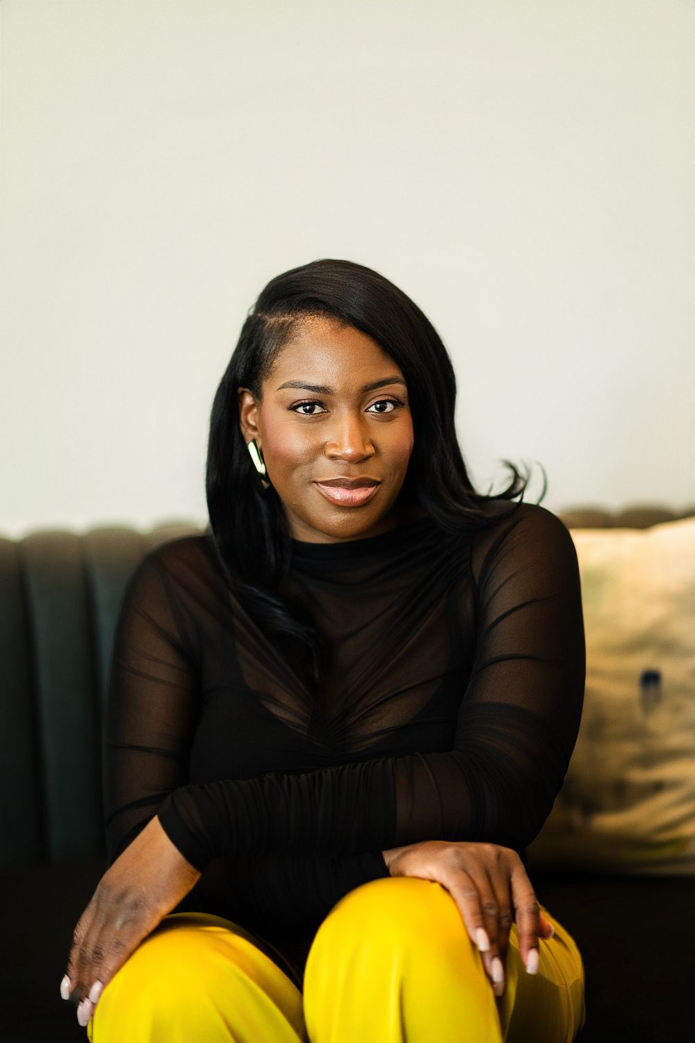 Smiling woman with long black hair in a sheer black top and bright yellow pants sits on a dark green sofa with her hands on her knees.