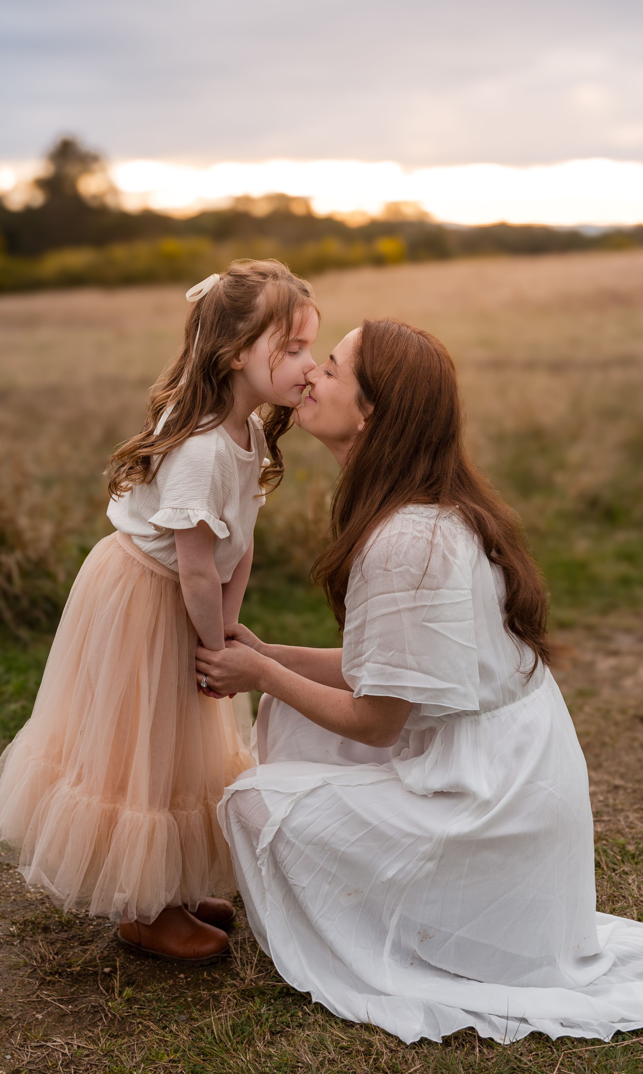 Mother and daughter touching noses in a sunlit Blue Mountains field, daughter in soft rose pink tunic, captured in authentic family moment.