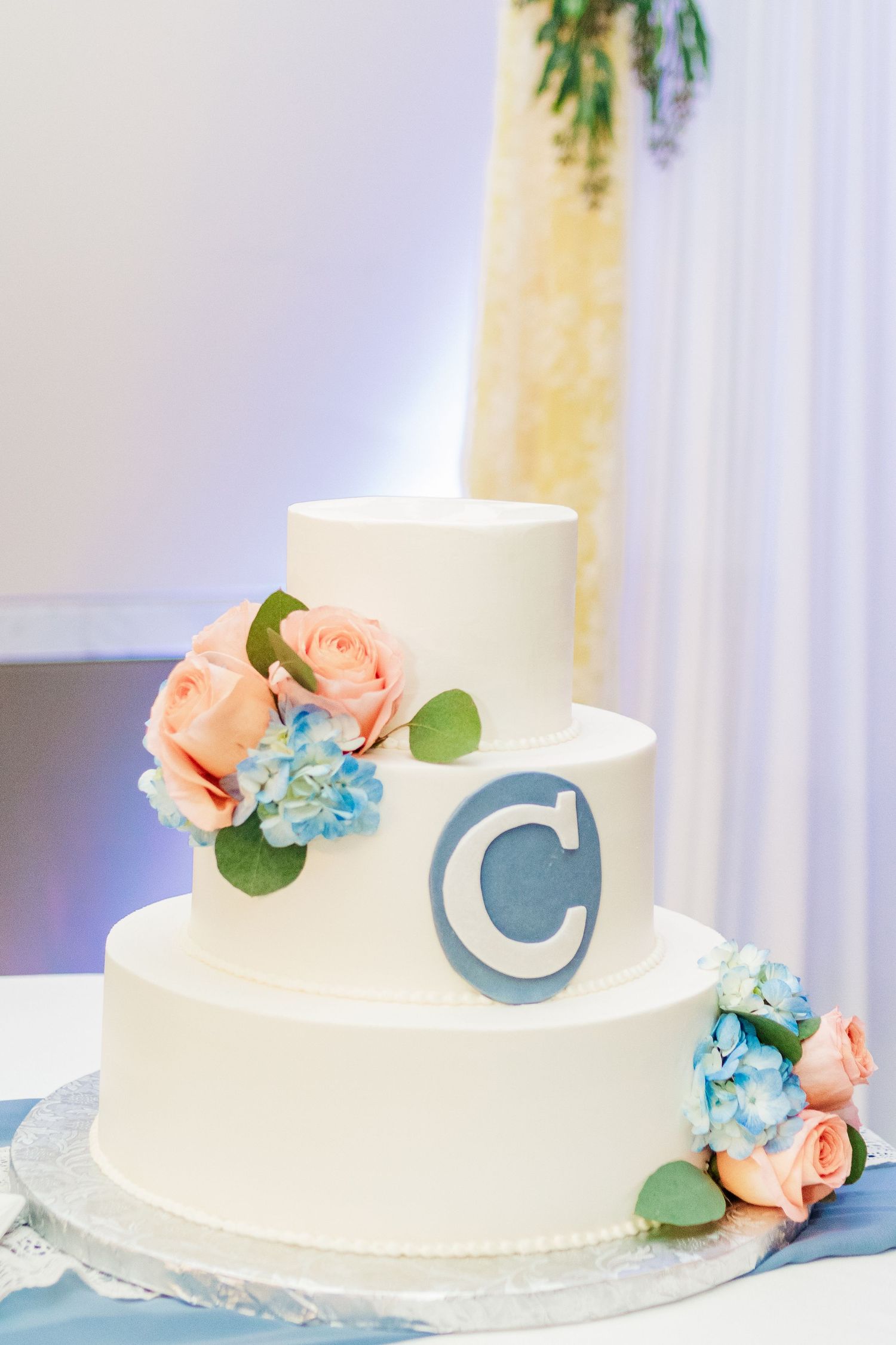 Three-tier white wedding cake decorated with peach roses and blue hydrangeas featuring a monogram C.