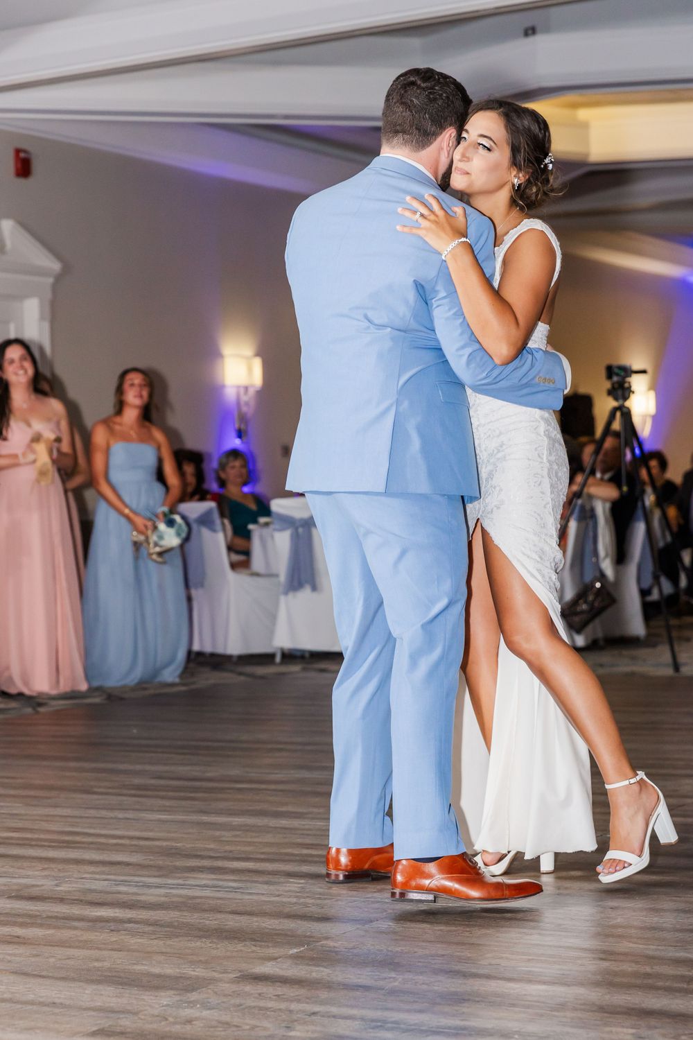 Couple dances intimately in a wedding reception hall with bridesmaids in pastel dresses watching in the background.