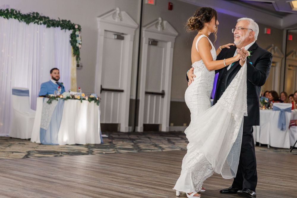 Wedding reception dance features white-gowned dancer with elderly partner as guests watch nearby.