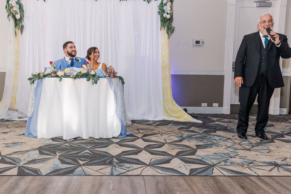 Wedding toast being given at reception while couple listens from decorated sweetheart table.