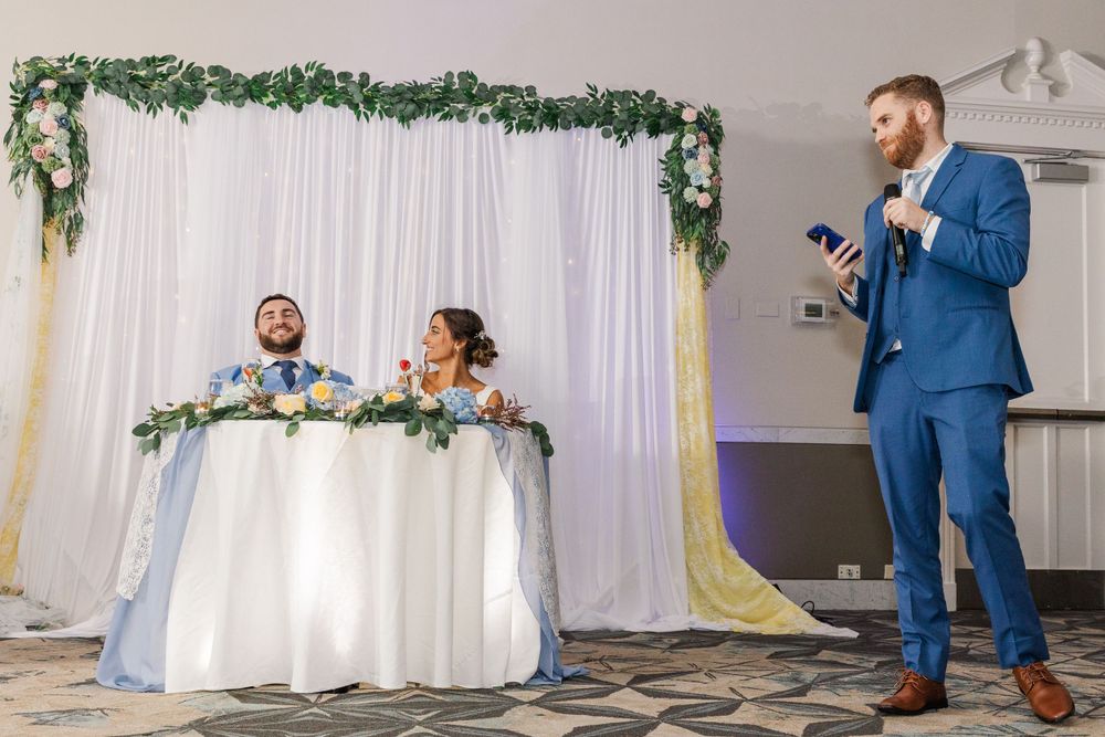 Wedding speech delivered by guest in blue suit as newlyweds listen from decorated head table.
