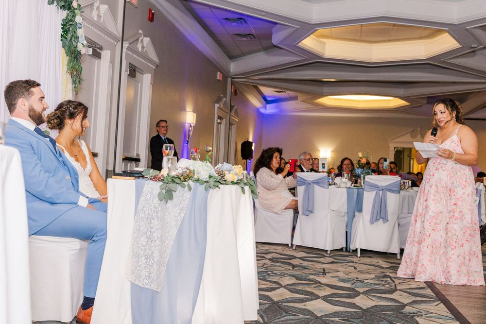 Wedding reception toast being given as couple listens from sweetheart table with floral decor.