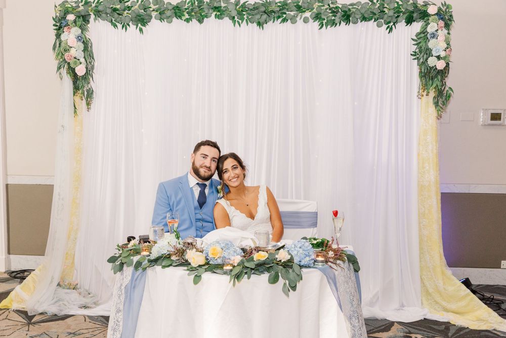 Newlyweds seated at elegant sweetheart table decorated with greenery and white backdrop.