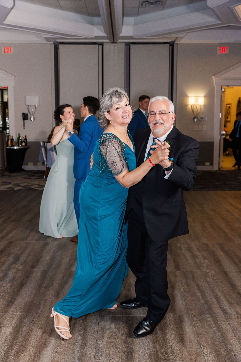 Couples dancing at a formal event on a wooden dance floor with one pair in teal and black formal attire.