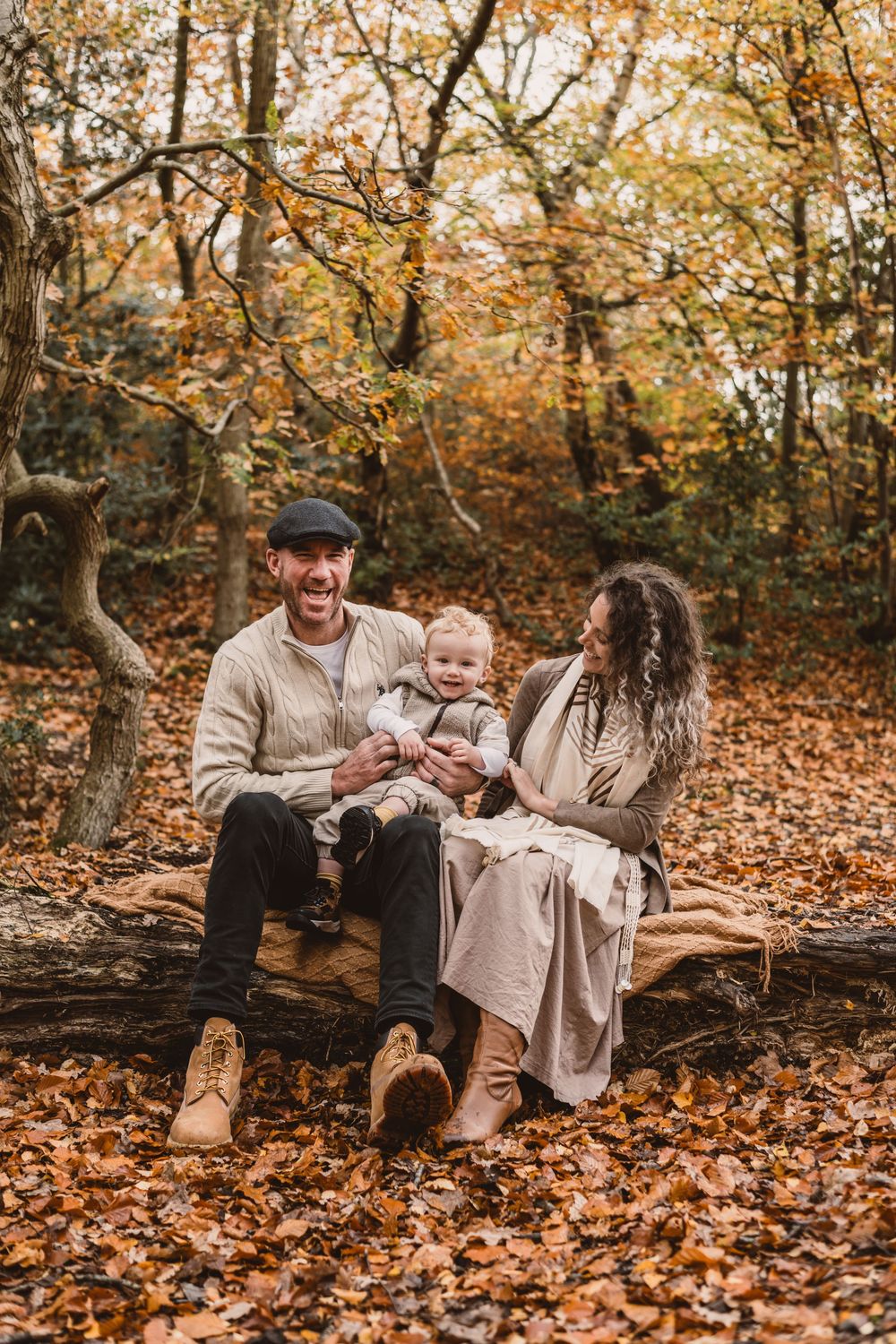 Family sitting together on fallen log surrounded by autumn leaves in forest setting.