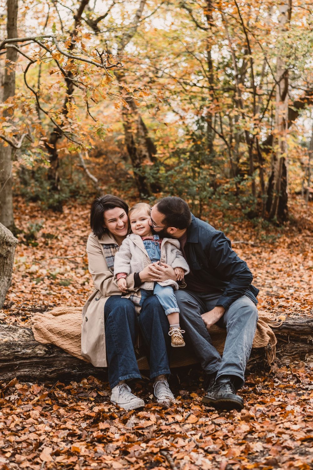 Family sitting together on fallen log surrounded by autumn leaves in forest setting.