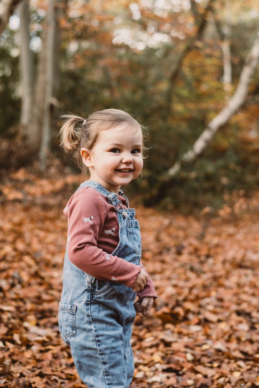 A young child in denim overalls and pink sweater plays joyfully in fallen autumn leaves on a crisp fall day.