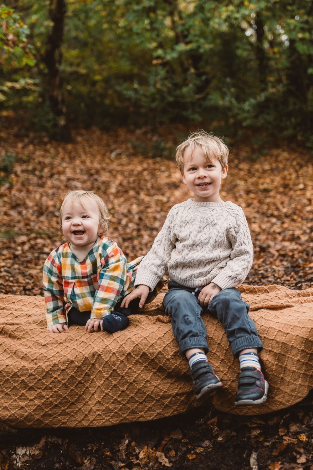 Children in autumn clothing sitting on a large brown blanket with fallen leaves in the background.