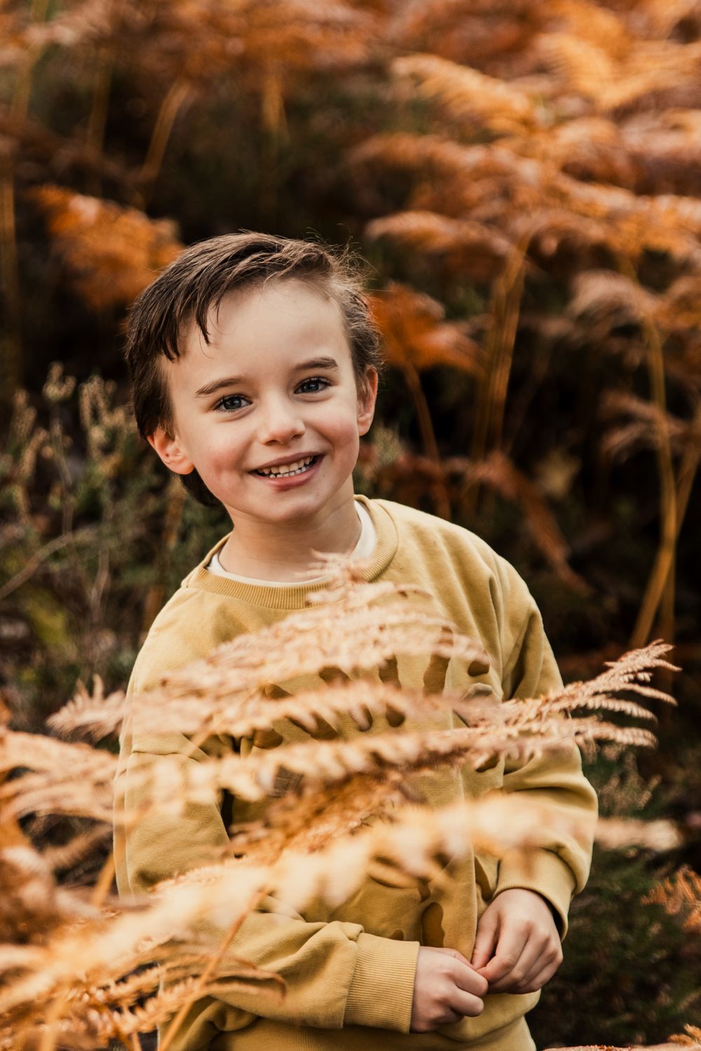 Child in yellow sweatshirt smiles brightly among golden autumn ferns.