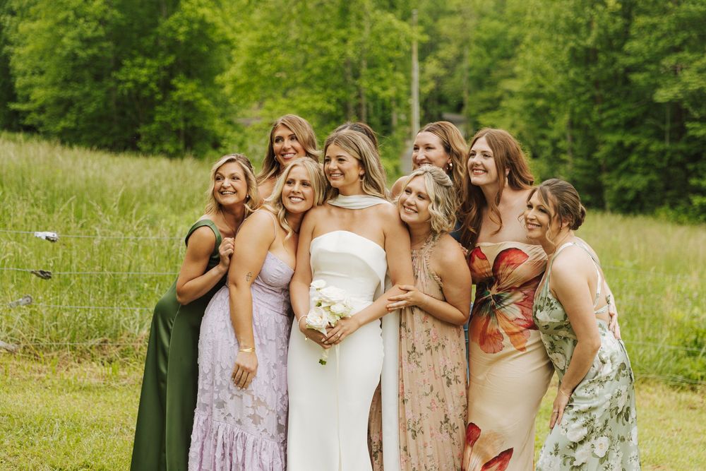 A group of women in formal dresses laugh together while standing in a grassy meadow surrounded by green trees.