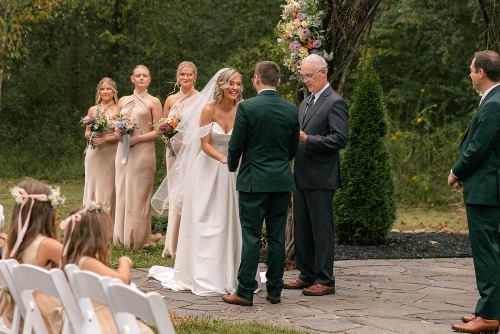 An outdoor wedding ceremony takes place on a stone patio with bridesmaids in beige dresses and white chairs for guests.