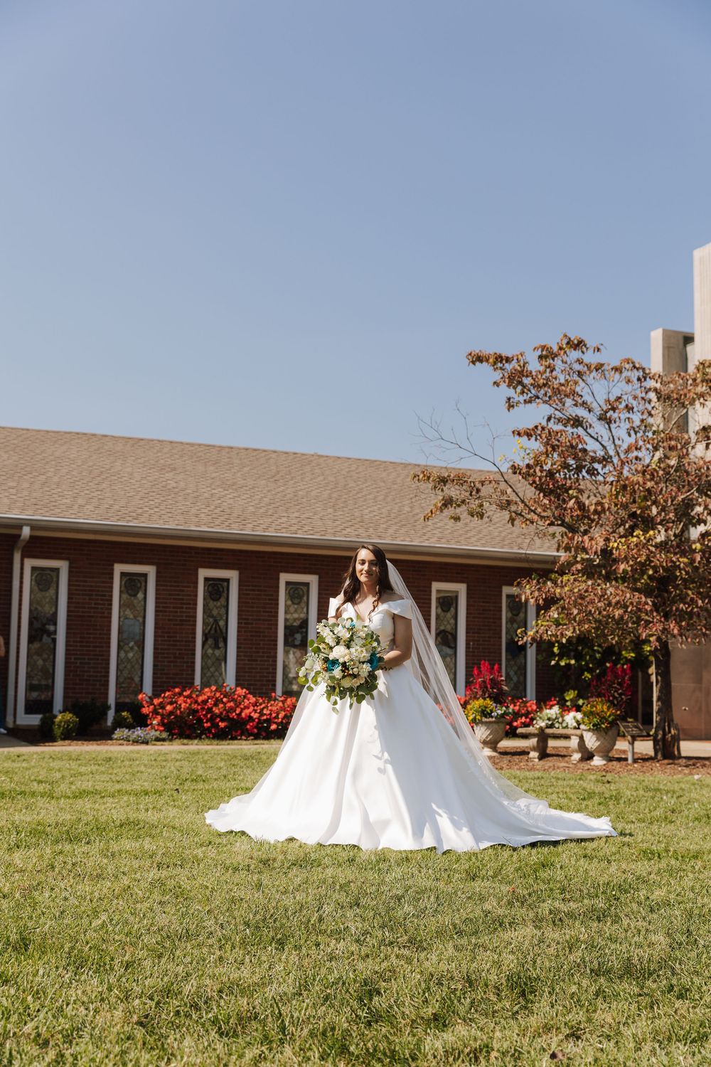 A bride in a flowing white gown holds a bouquet while standing on manicured lawn outside a church building.
