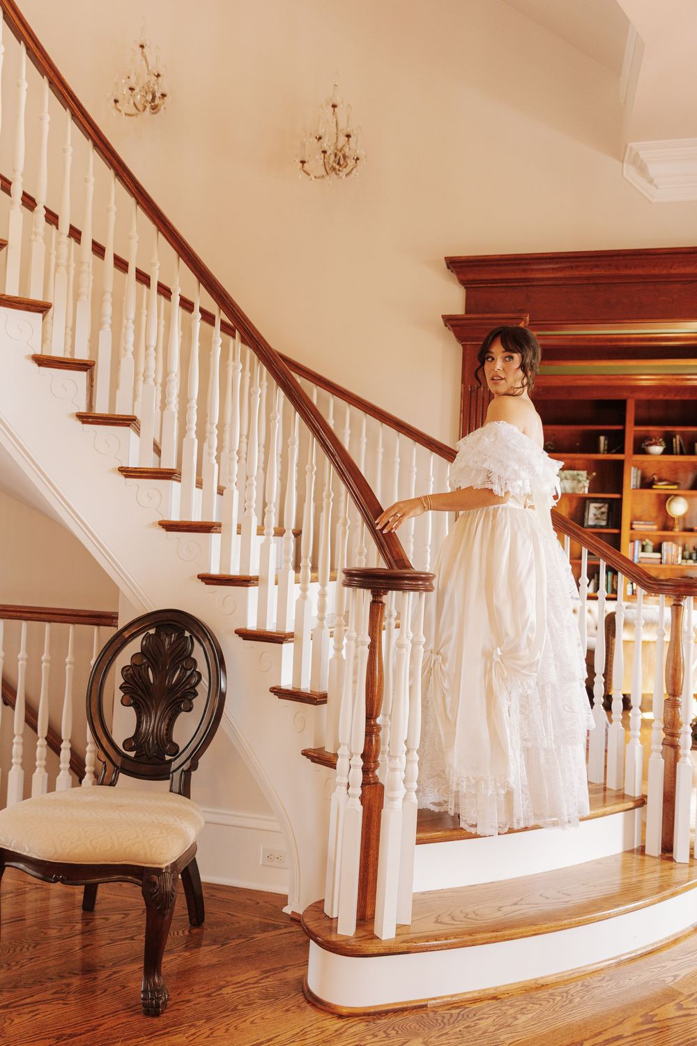 Classic staircase with white spindles and dark wood railings, featuring an antique chair and decorative chandeliers.