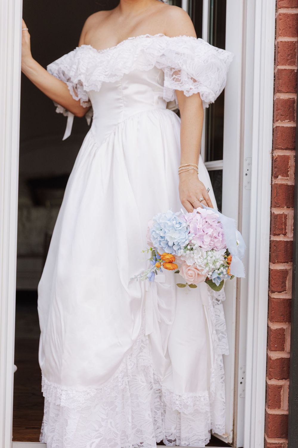 Elegant off-shoulder white wedding dress with lace trim and delicate pastel bouquet of blue and pink flowers.