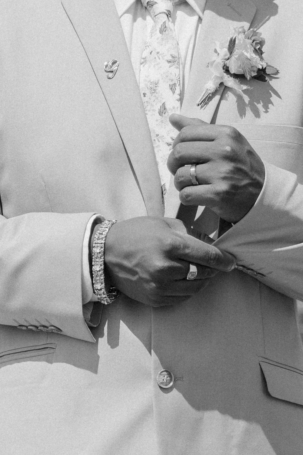 Close-up black and white photo showing hands adjusting a light suit jacket with a floral tie and diamond bracelet.