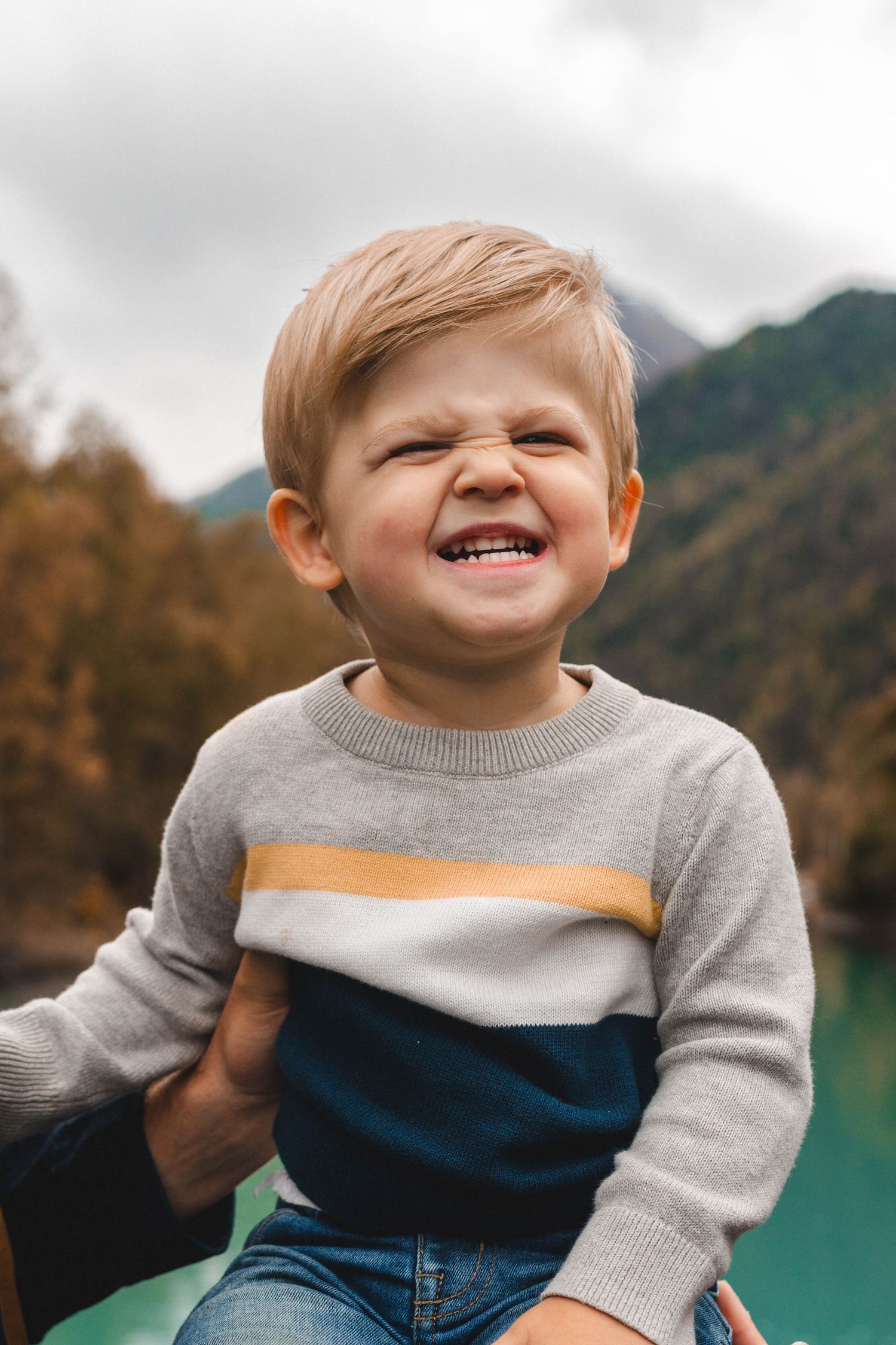 Adorable blonde toddler boy in striped sweater making a funny scrunched face near a mountain lake.