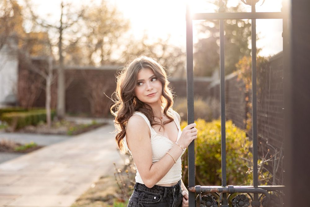 Young woman with long wavy hair poses by iron gate in golden sunlight wearing white top and dark jeans.