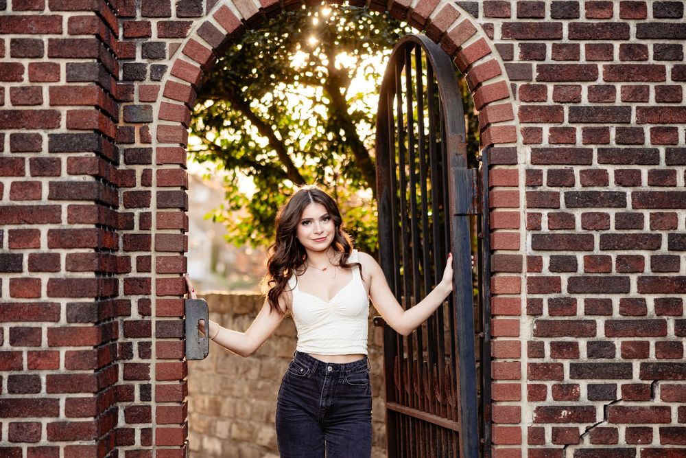 Young woman in white top and black jeans posing in a brick archway with iron gate and sunlit trees.