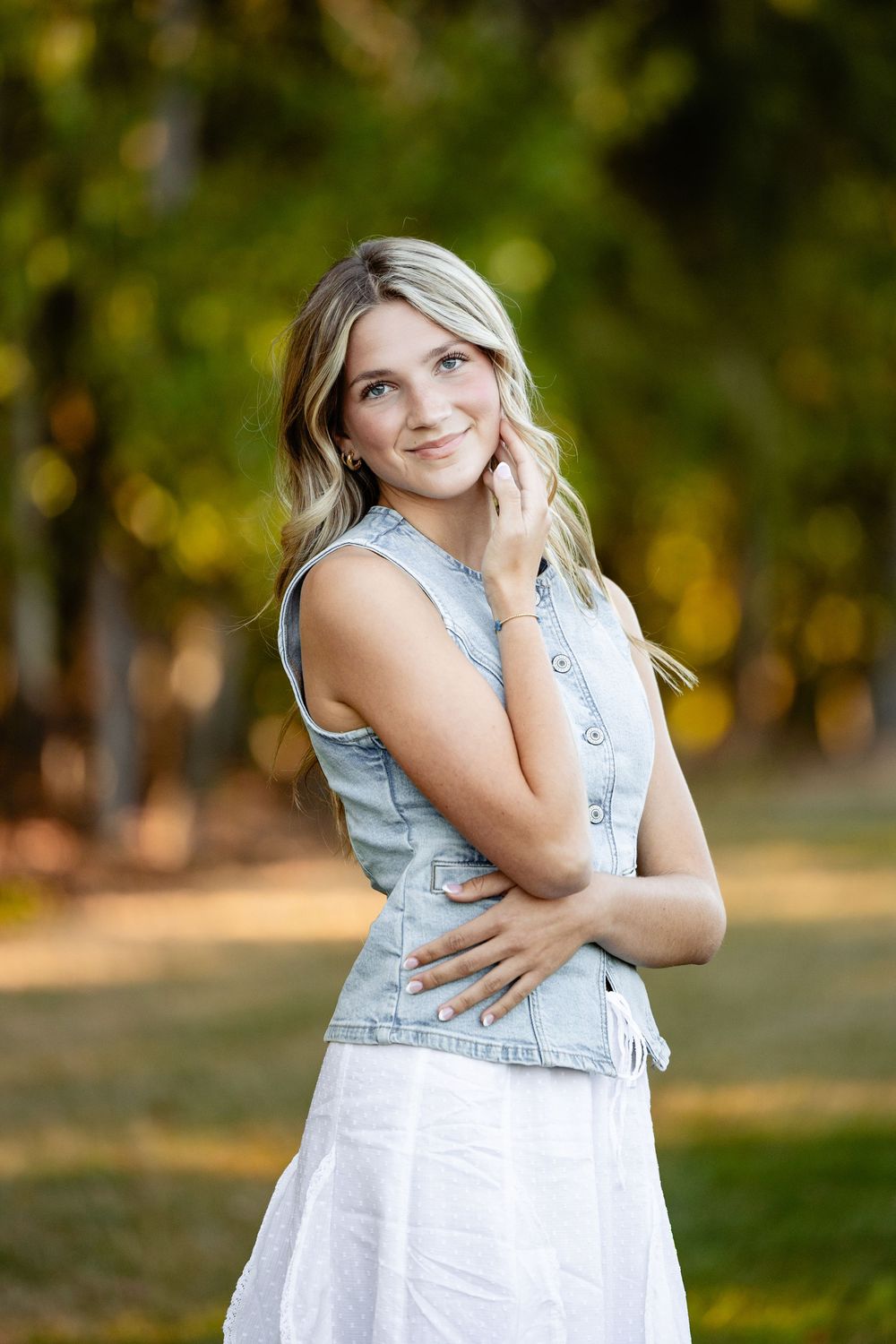 Young blonde woman in denim vest and white skirt poses outdoors with bokeh green trees in background.