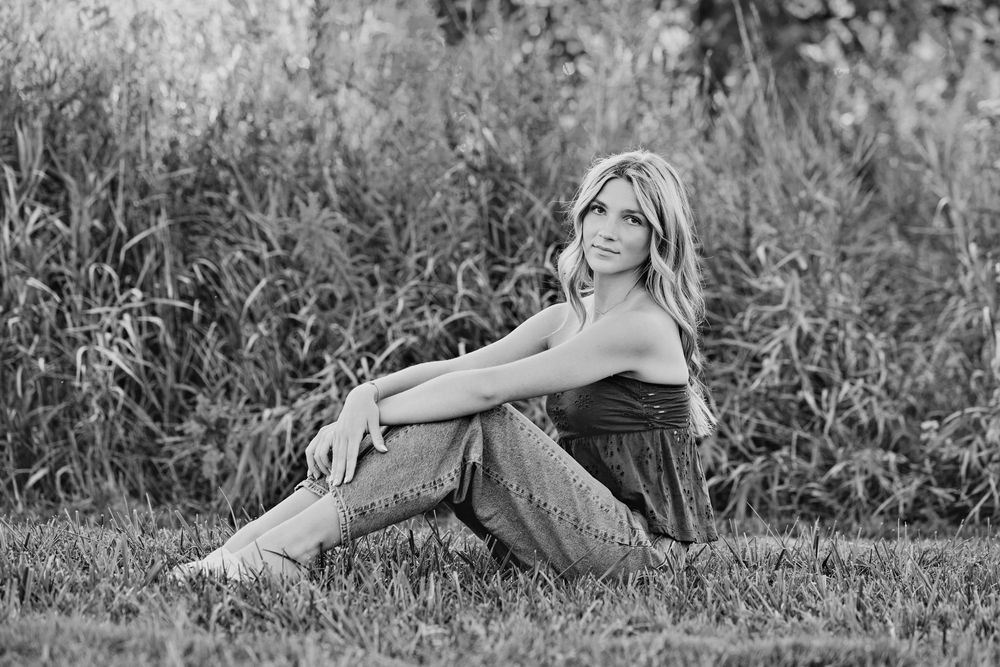 Black and white photo of a young blonde woman sitting on grass in jeans and a top, with tall reeds behind her.