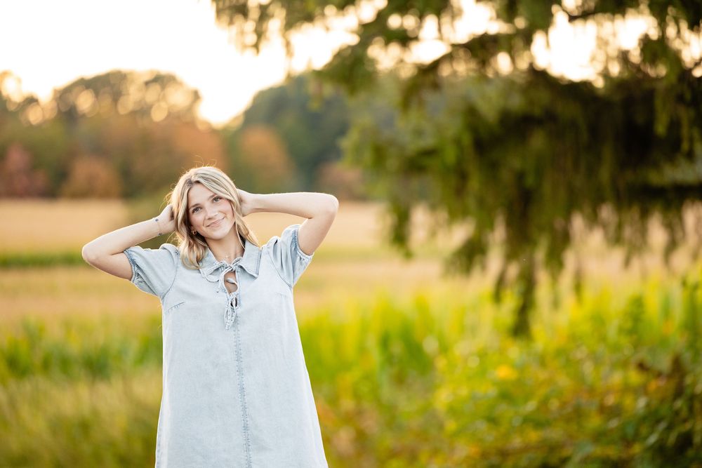 Young blonde woman in light blue dress posing outdoors in a sunlit field with lush green trees.