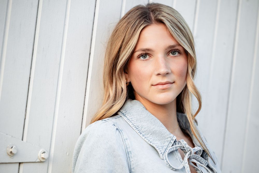 Young blonde woman in denim jacket leaning against white wooden wall in a professional portrait photo.