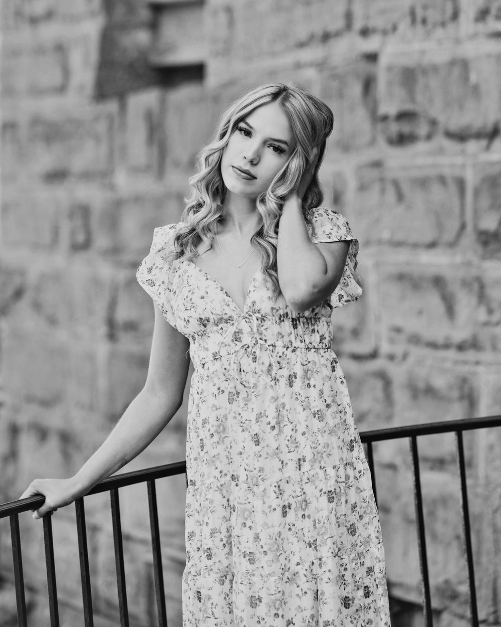 Black and white portrait of a young blonde woman in a floral dress leaning on a railing by a stone wall.