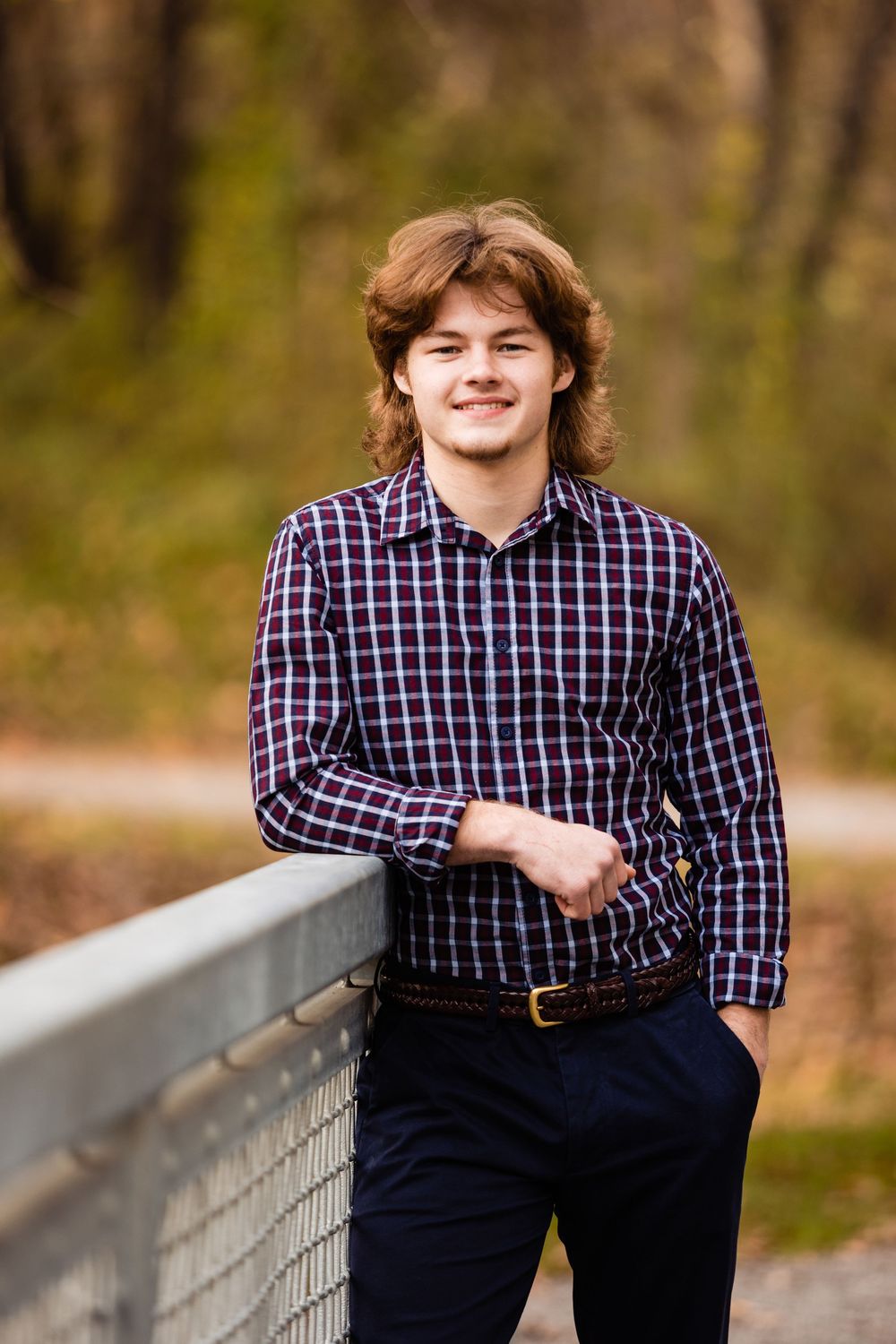 Young man with auburn hair smiling in plaid shirt, leaning on bridge railing in autumn park setting.